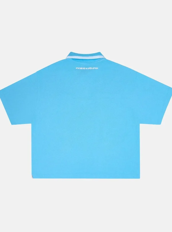Vista trasera del polo Cocky azul de Cold Culture, con diseño liso, logotipo blanco bajo el cuello y mangas cortas de corte amplio.