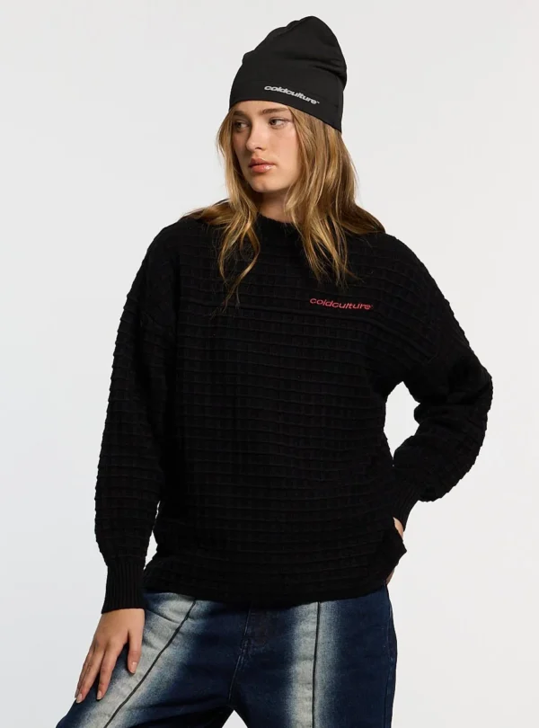 Modelo con jersey negro Crunchy Waffle Knit de Cold Culture, con textura de punto tipo gofre, mangas holgadas y pequeño logotipo rojo en el pecho.