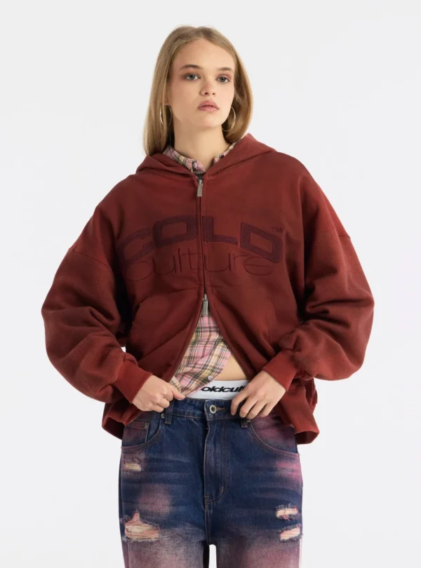 Modelo con sudadera roja Dust 55 de Cold Culture, con logotipo frontal, mangas amplias y cremallera de doble cursor parcialmente abierta.