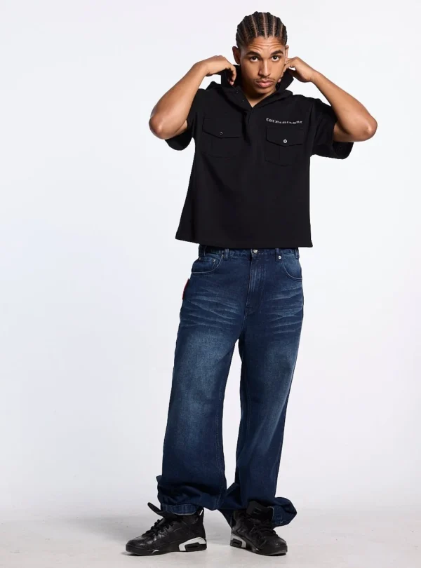 Modelo de cuerpo entero vistiendo el Gilded Polo negro de Cold Culture con vaqueros anchos azules y sujetando el cuello del polo.