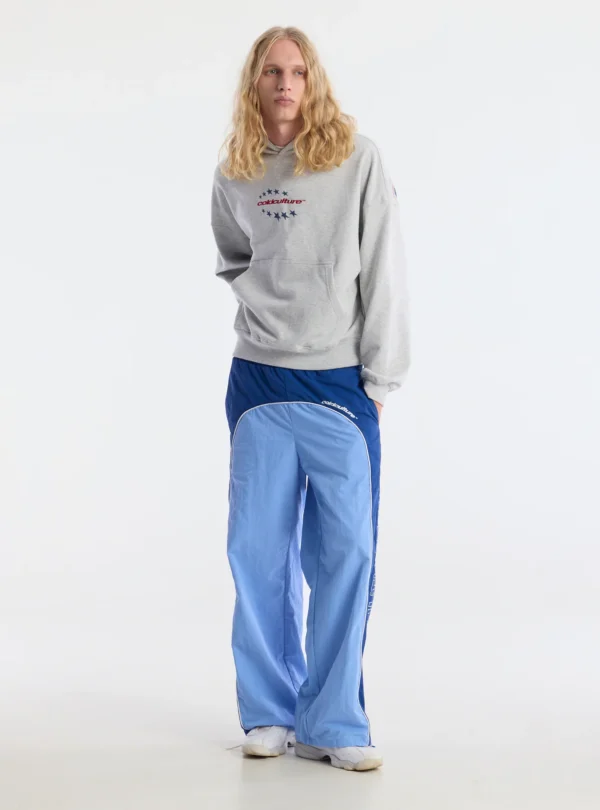 Modelo con pantalones deportivos Overcut de Cold Culture en azul, de corte ancho y paneles bicolor, combinados con una sudadera gris con capucha y zapatillas blancas.