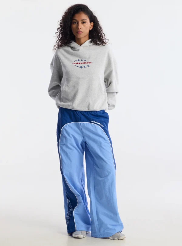 Modelo con pantalones deportivos Overcut de Cold Culture en azul claro y azul oscuro, con perneras anchas, ribetes blancos y estampado lateral, combinados con una sudadera gris y zapatillas deportivas.