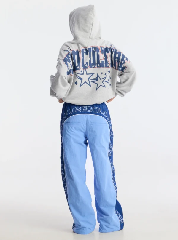Vista trasera de una modelo con pantalones deportivos Overcut de Cold Culture en azul, con paneles bicolor, estampado CULTURE COLD en la cintura y los laterales, y perneras de corte ancho.