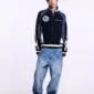 Modelo con jersey Pole Knit Navy de Cold Culture, con cremallera frontal, escudo en el pecho y mangas con rayas, combinado con vaqueros anchos azules.