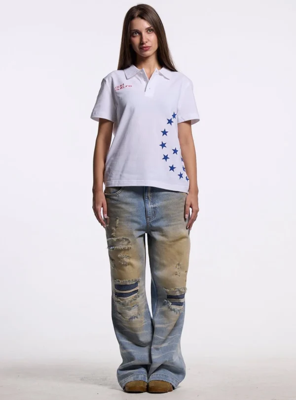 Modelo femenina con polo blanco Starttrail de Cold Culture, logotipo rojo en el pecho, estrellas azules y vaqueros desgastados.