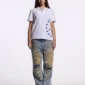 Modelo femenina con polo blanco Starttrail de Cold Culture, logotipo rojo en el pecho, estrellas azules y vaqueros desgastados.
