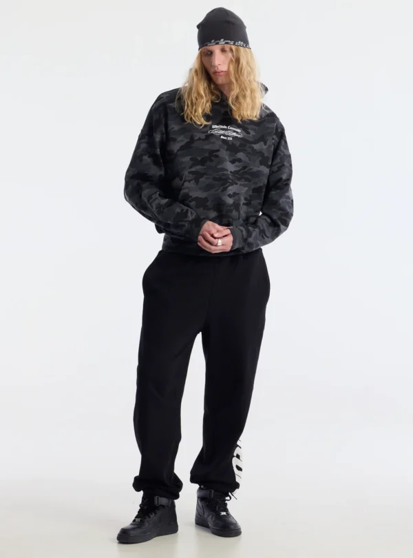 Modelo con pantalones deportivos negros Vertical de Cold Culture, combinados con sudadera de camuflaje oscuro y zapatillas negras.