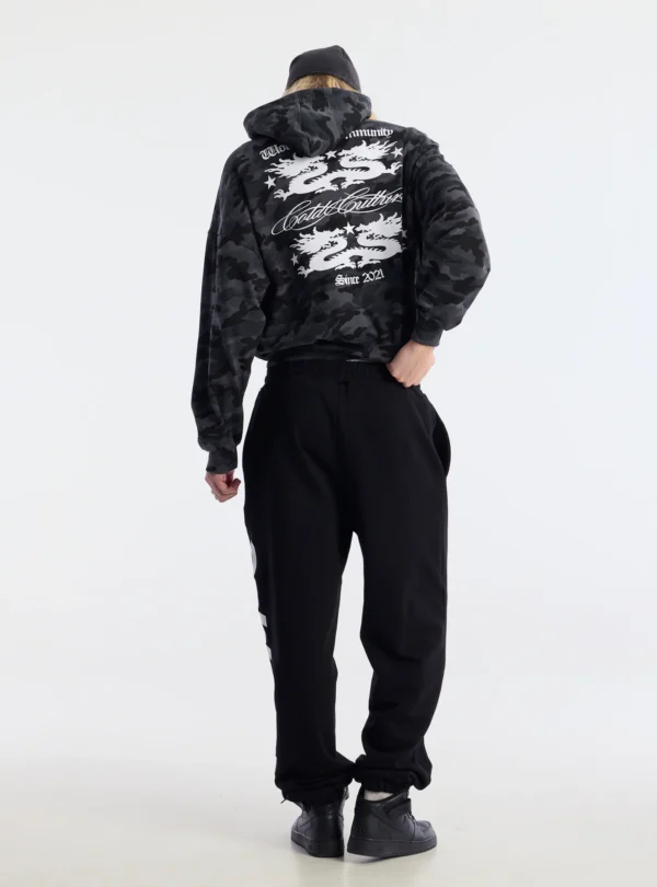 Modelo de espaldas con pantalones deportivos Vertical Black de Cold Culture, de corte holgado y puños elásticos, combinados con sudadera de camuflaje.