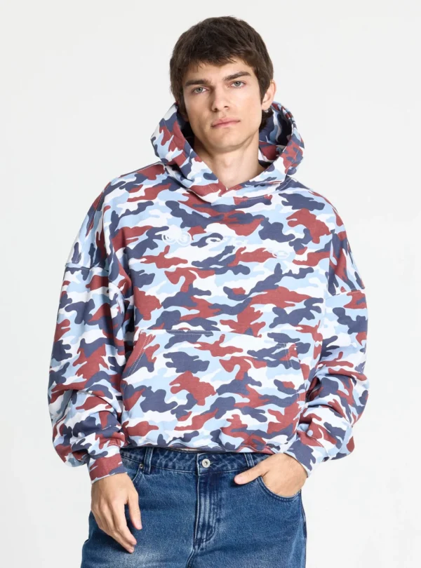Cold Culture Vertical Sudadera – Camo – Detalle frontal en modelo Vista frontal de la sudadera Vertical de Cold Culture en camuflaje azul, rojo y blanco, llevada por un modelo, con capucha, hombros caídos y bolsillo canguro.
