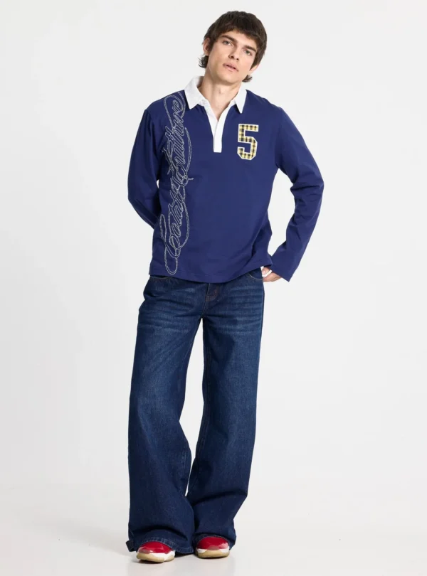 Modelo con el polo Windsor Longsleeve Polo Navy de Cold Culture, azul marino con cuello blanco y número 5, combinado con vaqueros anchos oscuros.